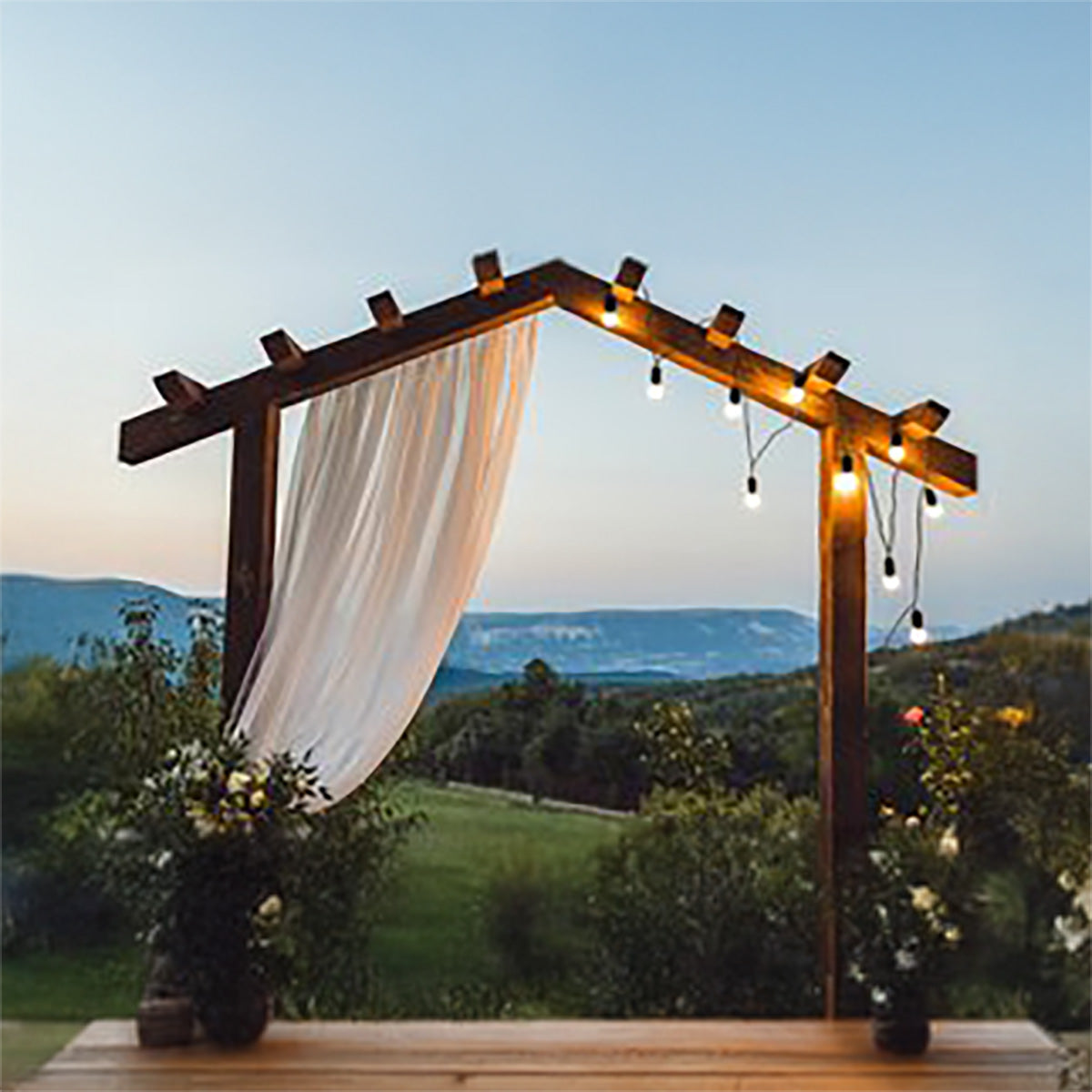 Wooden arch with white curtain and string lights against a scenic background