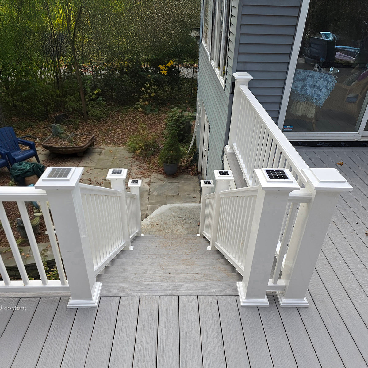 Gray deck with white railings leading to a house, surrounded by greenery.