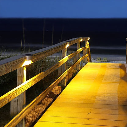 Wooden deck with railing illuminated by lights at night, overlooking a beach.